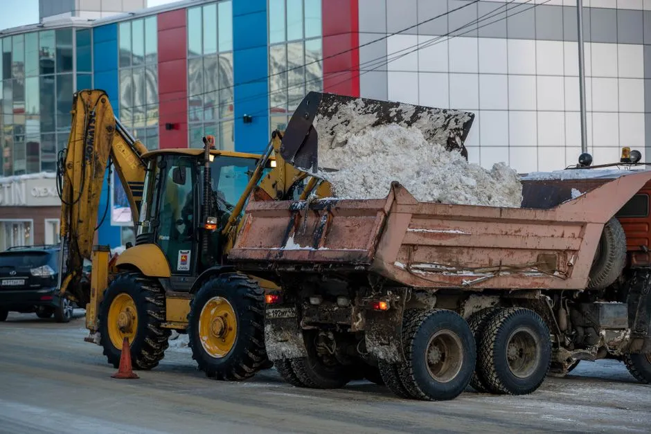 A backhoe and dump truck work together to clear snow in a city setting.